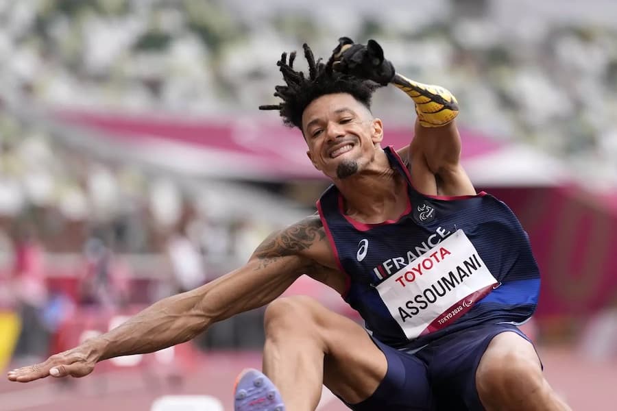 Athlete with dreadlocks mid-jump, with an arm prosthesis, during a Paralympic jumping competition